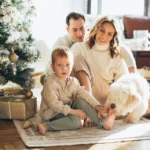Родина з дитиною сидить на підлозі біля прикрашеної різдвяної ялинки й гладить пухнастого білого собаку в теплому світлі з вікна